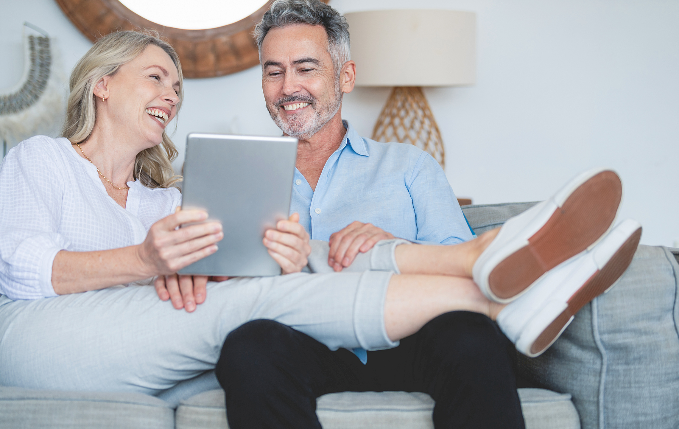 Ehepaar sitzt glücklich auf dem Sofa, sie legt die Beine  hoch und beide schauen in ein Tablet